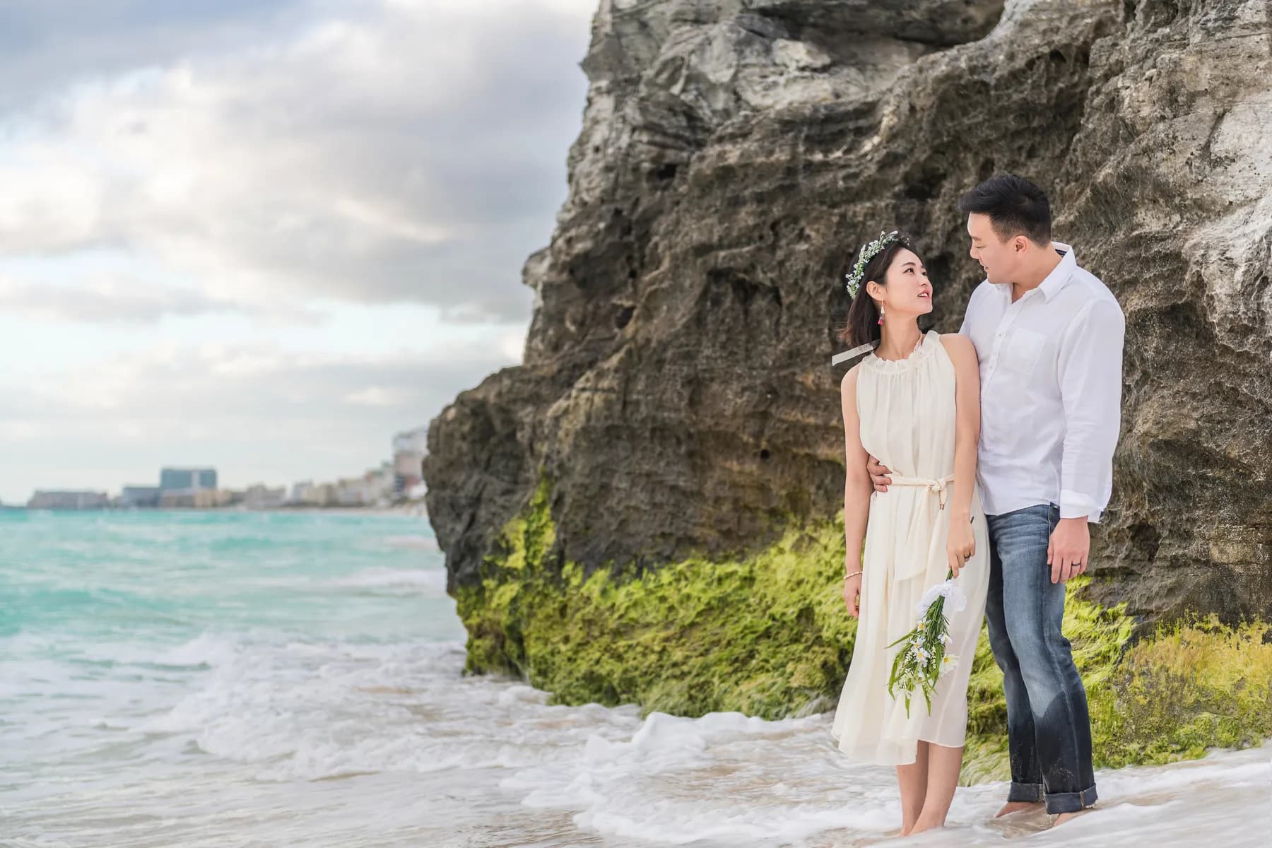 Couple standing by the waves beneath a coastal rock